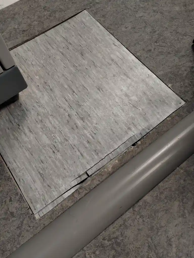 A close-up photo shows a square, gray, wood-grain patterned floor tile that is visibly cracked and lifted at its edges, set…