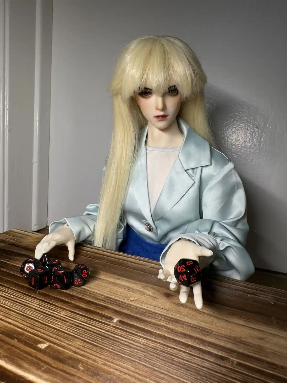 A doll with long blonde hair and a light blue satin jacket sits at a wooden table, holding one black die with red numbers…