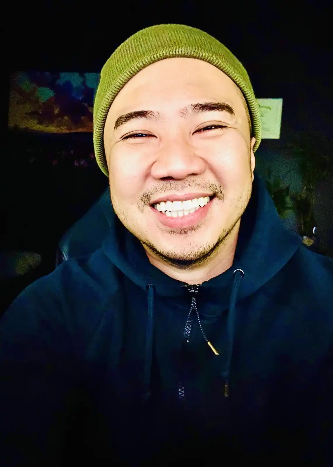 A man with a shaved head wearing a green beanie and a dark blue zip-up hoodie smiles broadly with his eyes crinkled shut…