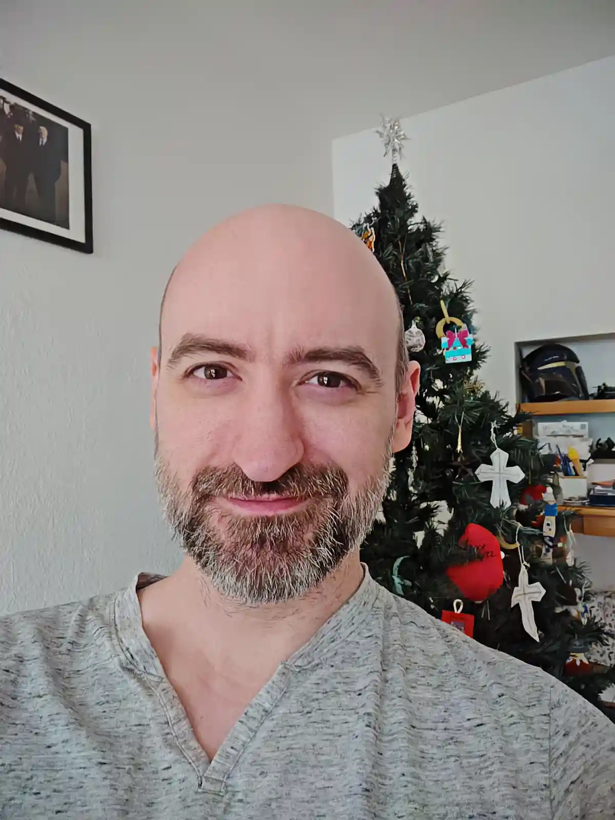 A bald man with a graying beard and a subtle, knowing smirk looks at the camera, standing in front of a decorated christmas…