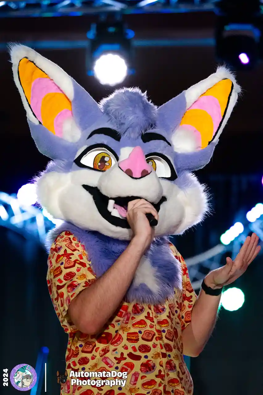 A person in a large, colorful blue and white fursuit with pink and yellow inner ears is singing into a microphone on a stage…