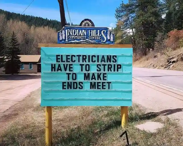 A turquoise wooden sign with black lettering reads 'ELECTRICIANS HAVE TO STRIP TO MAKE ENDS MEET', mounted on yellow posts…
