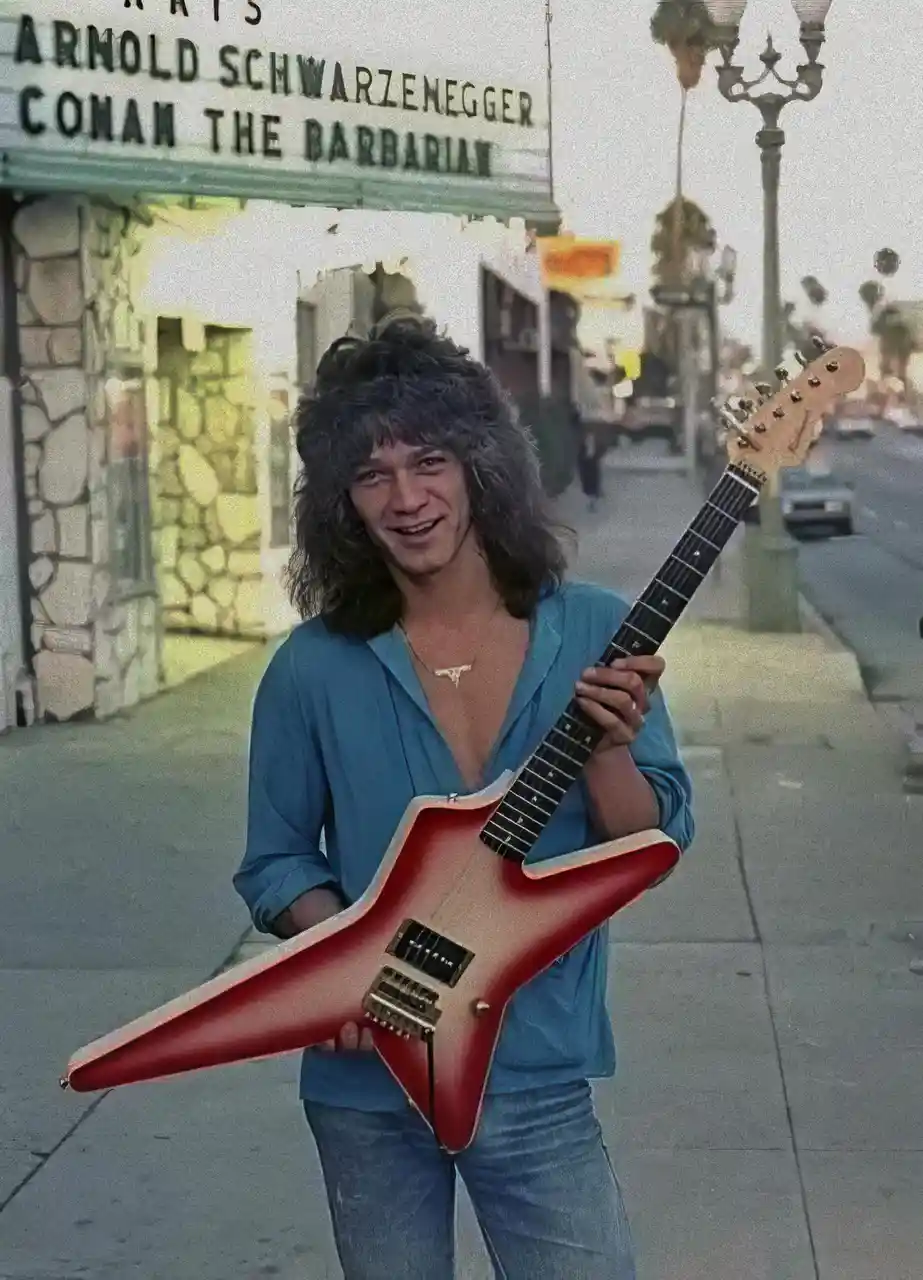 A young man with long, shaggy hair and a blue shirt holds a red star-shaped electric guitar while standing on a sidewalk…