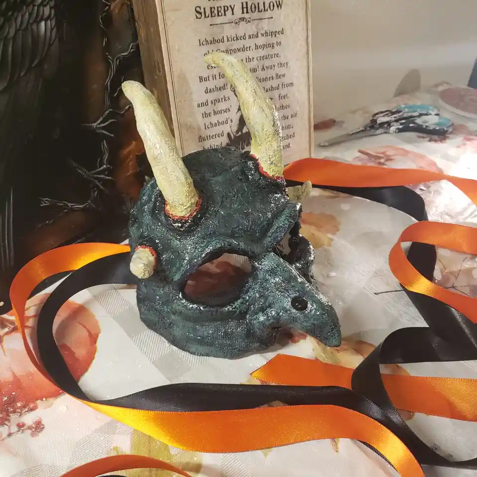 A handmade, dark green, textured devil mask with two prominent yellow horns and red accents sits on a table surrounded by…