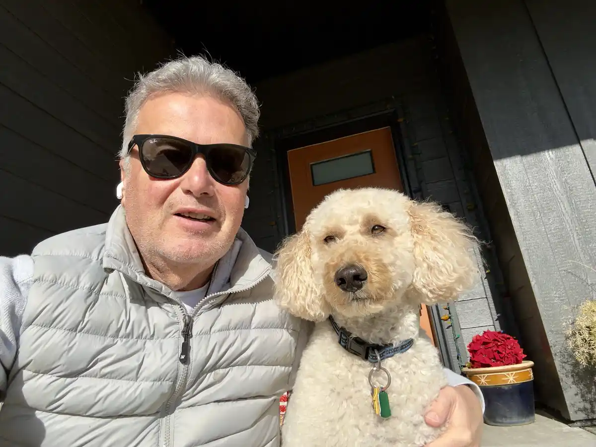 A man with gray hair, wearing sunglasses and a light gray puffer vest, holds a fluffy, light-colored poodle-like dog in…