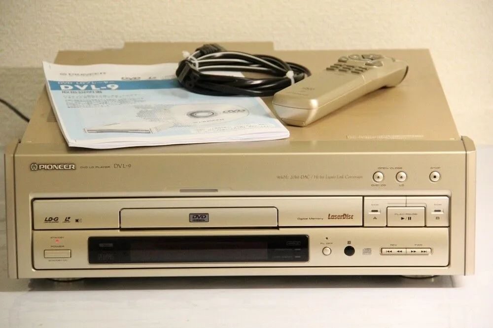 A beige Pioneer DVL-9 DVD player sits on a white surface with its manual, a coiled cable, and a matching remote on top. The…