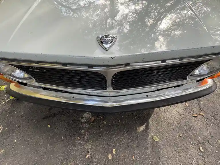 A close-up photo of the front of a light gray Volvo car, showing its chrome grille, headlights, and hood emblem. The car…