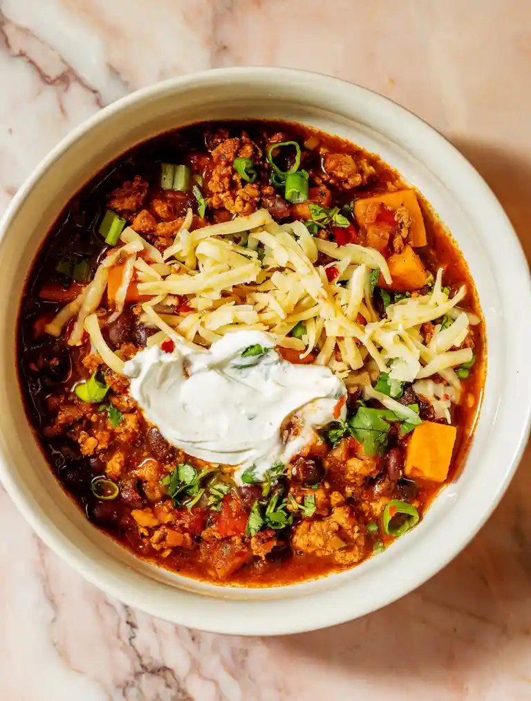 A top-down photo of a white bowl filled with rich, dark chili topped with shredded cheese, a dollop of sour cream, chopped…