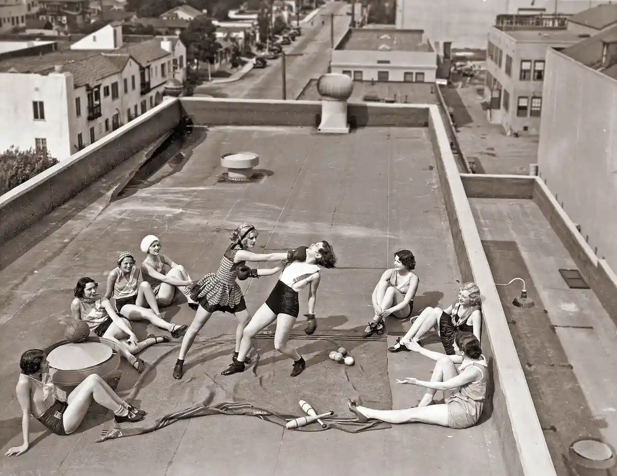 A vintage black-and-white photo shows eight women on a rooftop, two in boxing stances mid-fight while six others sit around…
