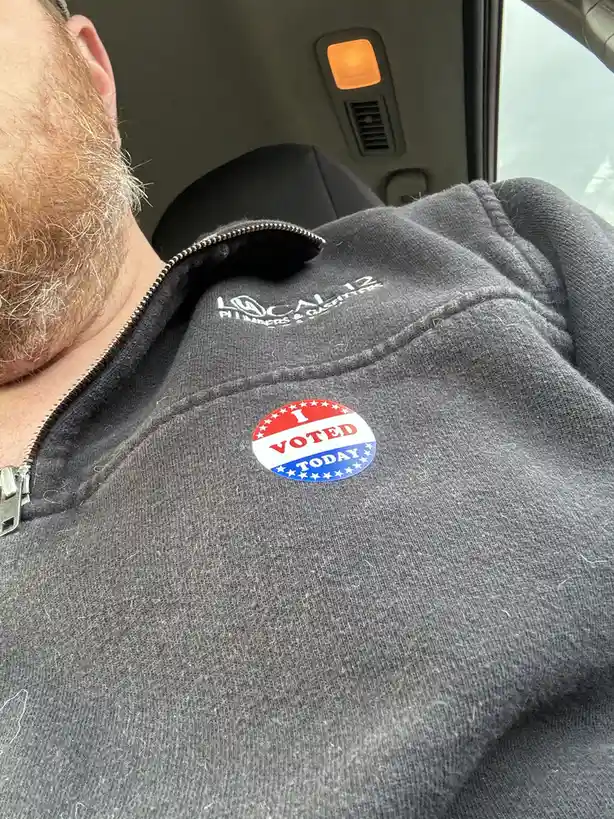A close-up photo shows the back of a man's head and his grey zip-up hoodie while he's sitting in a car. A circular 'I VOTED…