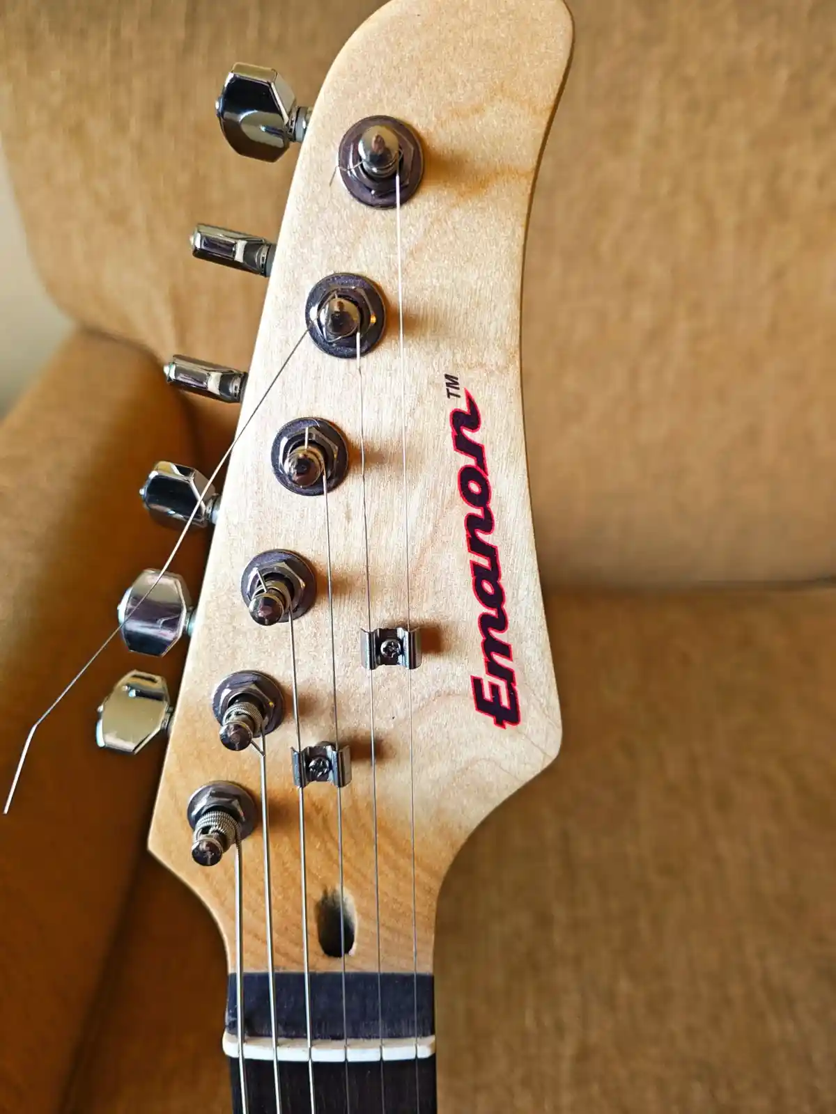 A close-up photo of an Emanon guitar headstock, showing the brand name in red lettering, six tuning pegs, and strings…