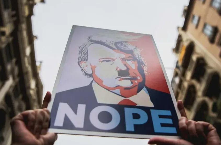 Two hands hold up a stylized poster featuring a portrait of Donald Trump with a stern expression, set against a red and blue…