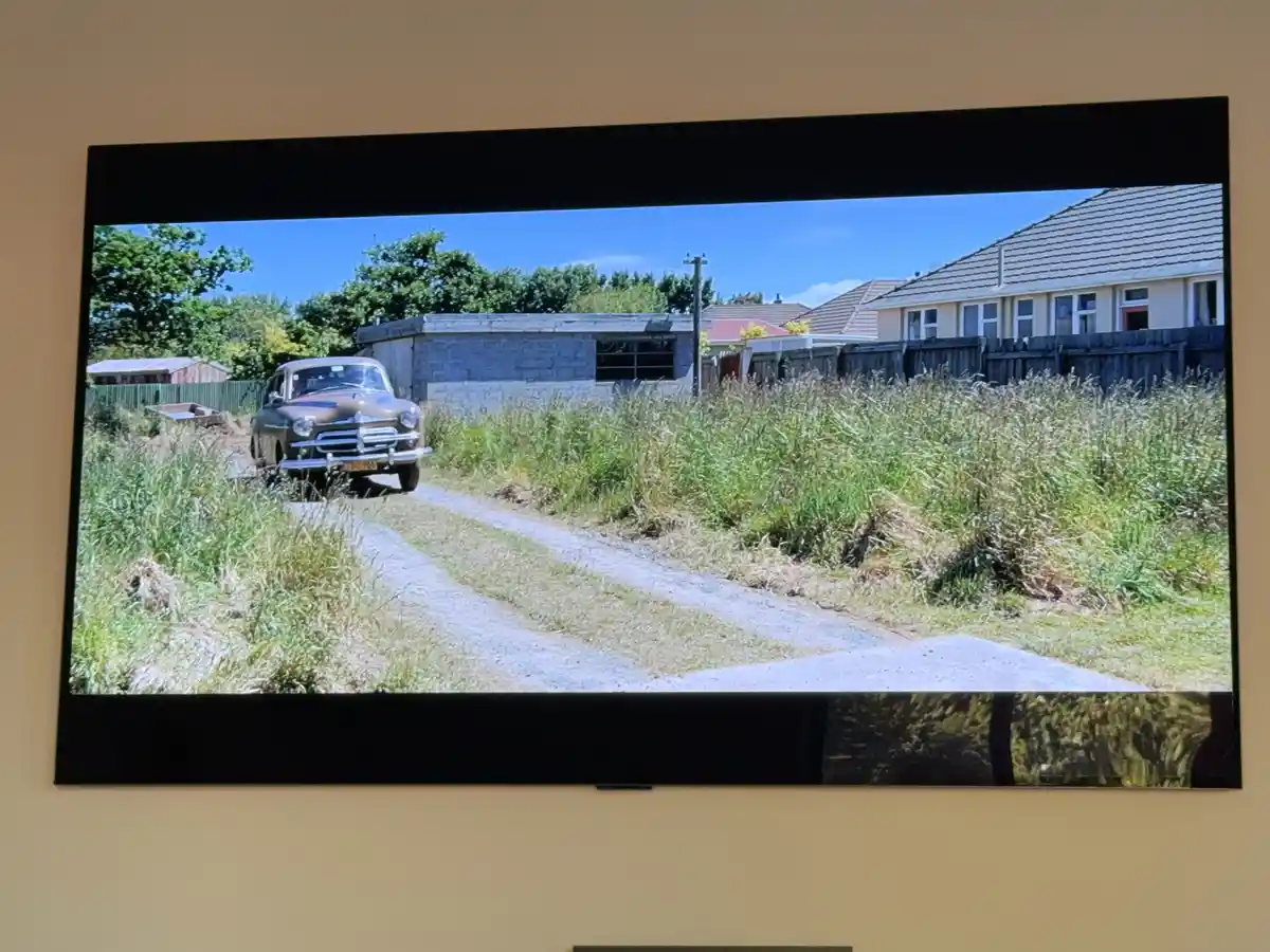 A television screen displays a scene of a vintage brown car driving down a gravel road surrounded by tall grass, with houses…