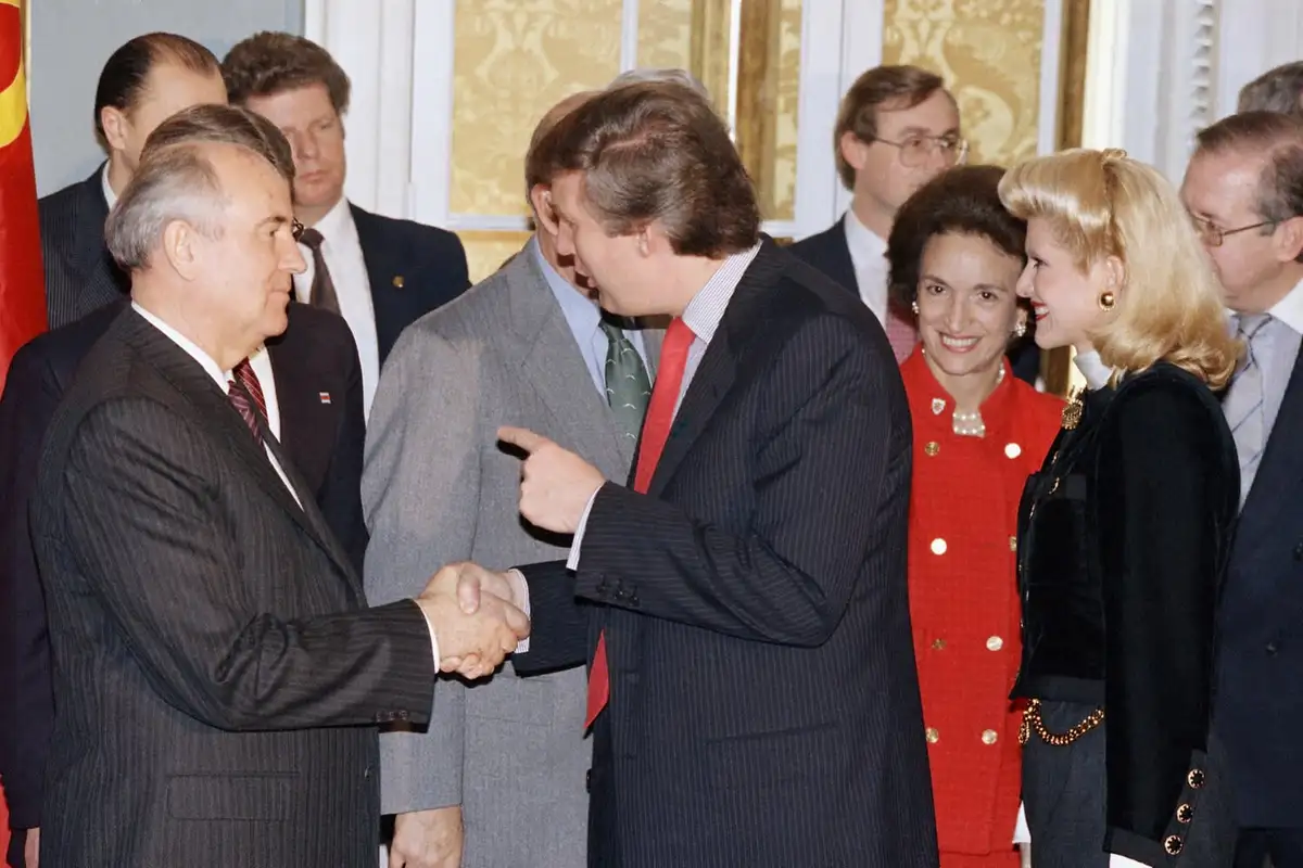a man in a dark pinstripe suit shakes hands with another man while pointing at him, with a group of formally dressed people…