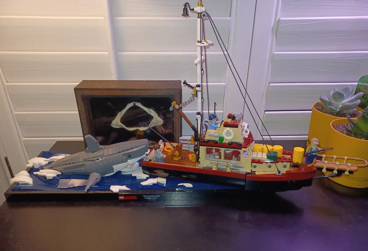 A LEGO Jaws set depicts a small red and beige boat with minifigures, facing a large grey LEGO shark emerging from blue LEGO…