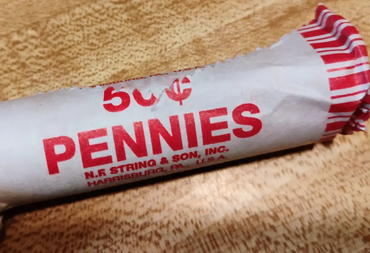 A close-up photo shows a roll of U.S. pennies wrapped in paper printed with '50¢ PENNIES' and 'N.F STRING & SON, INC…