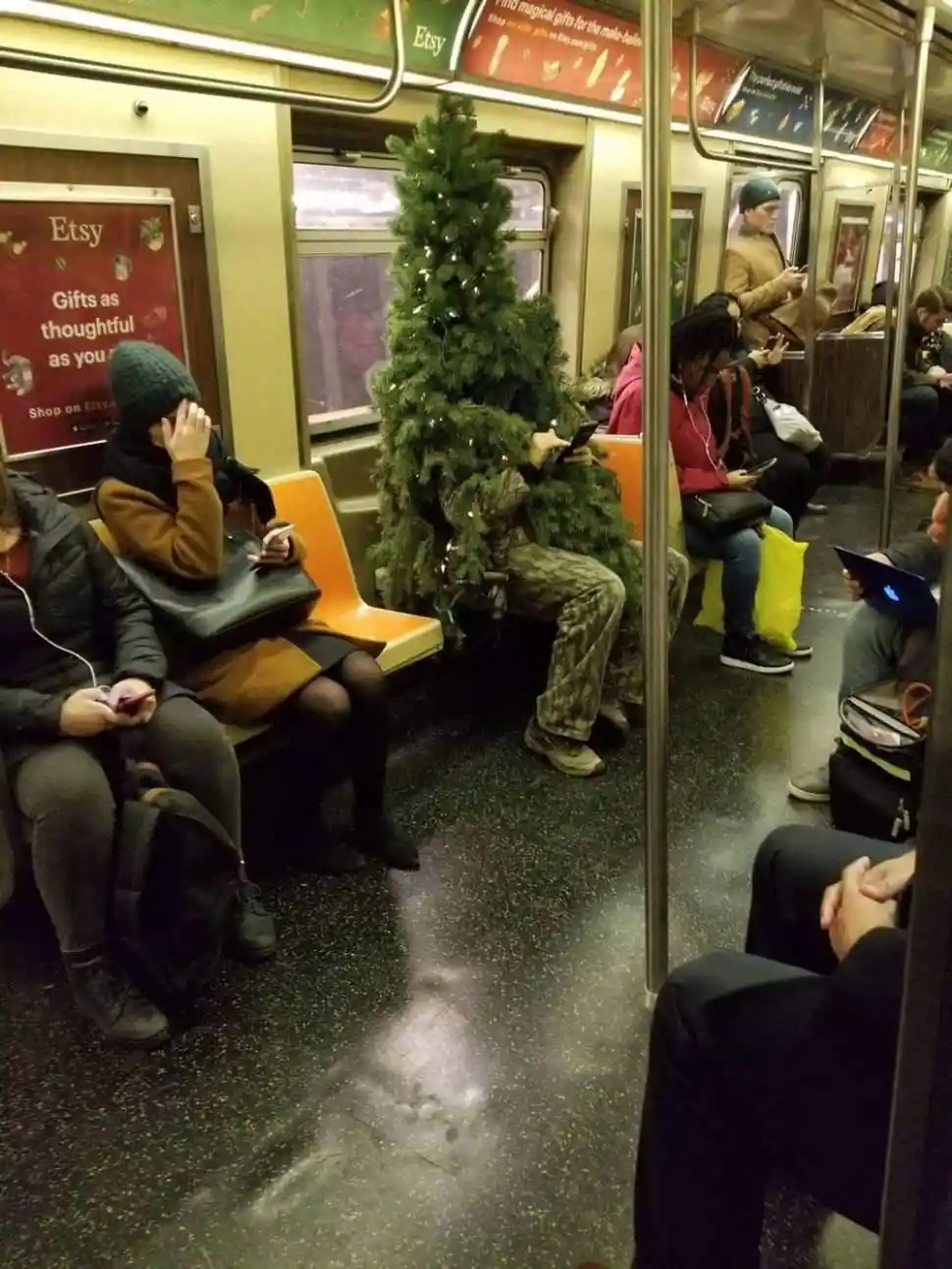 A person sitting on a subway train is completely covered by a small, decorated christmas tree, with only their legs and feet…