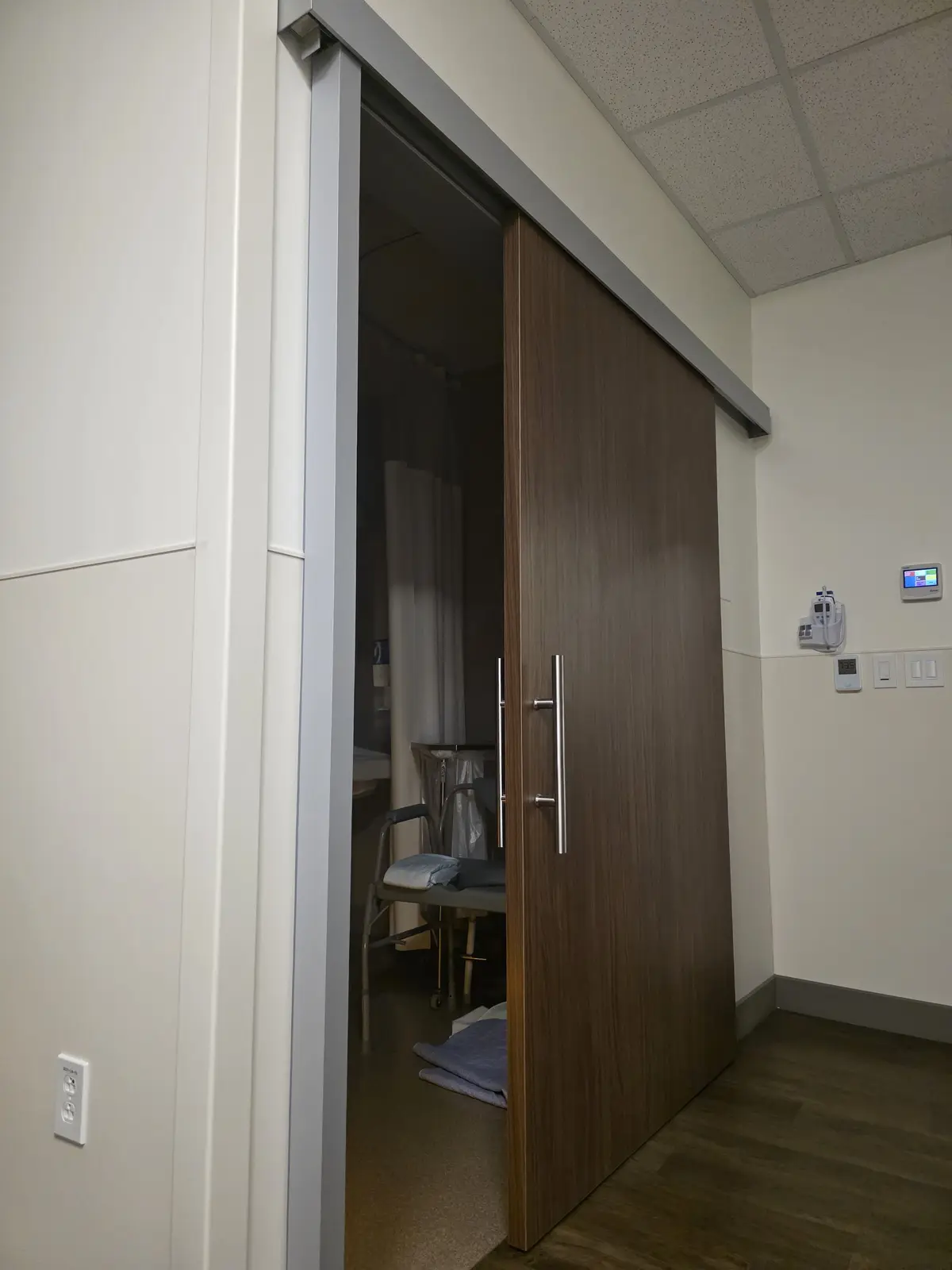 A partially open sliding door in a medical facility reveals a hospital room with a wheelchair, curtain, and medical…