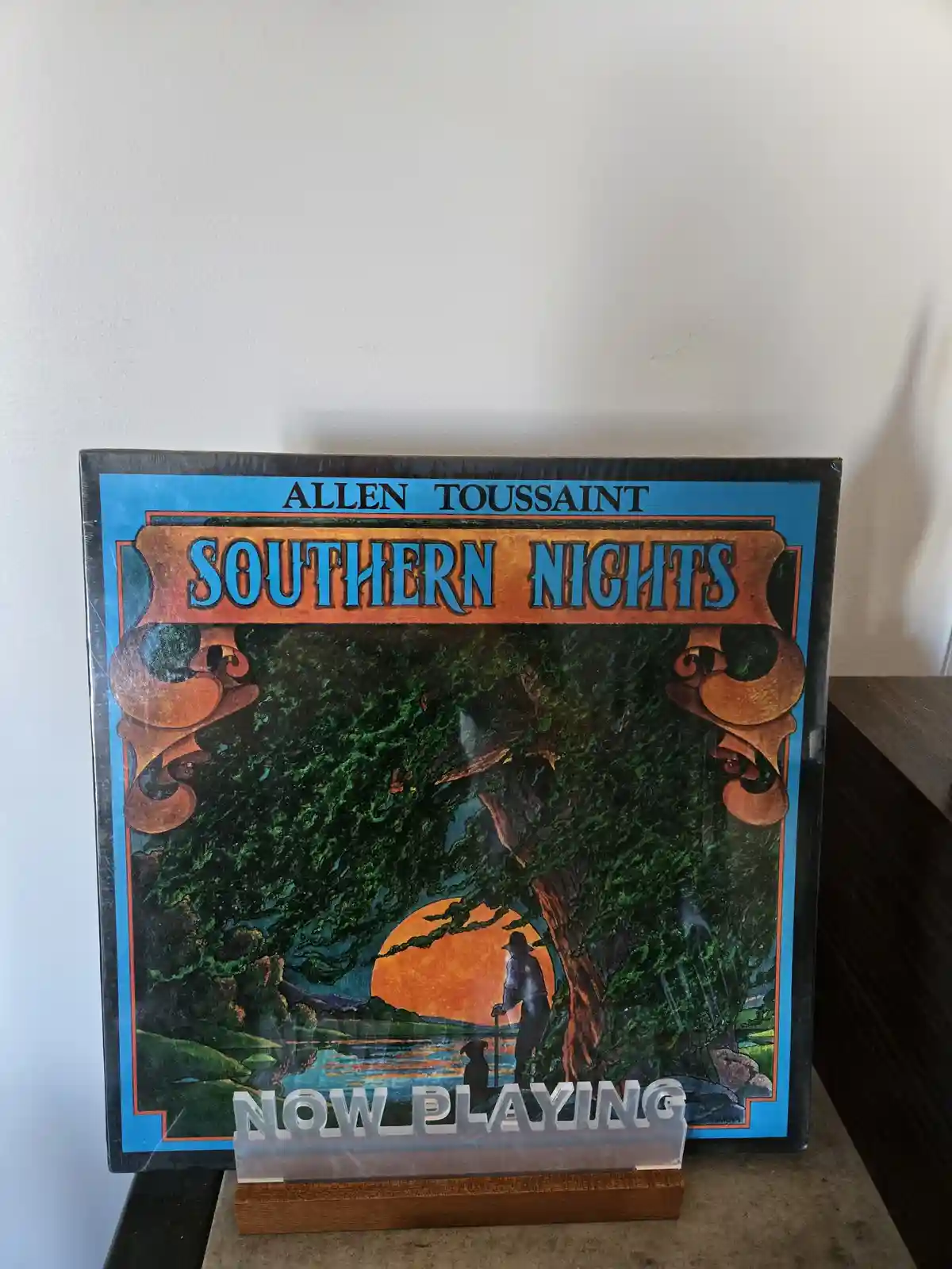 A vinyl record album cover for Allen Toussaint's 'Southern Nights' is displayed upright on a wooden stand, featuring an…