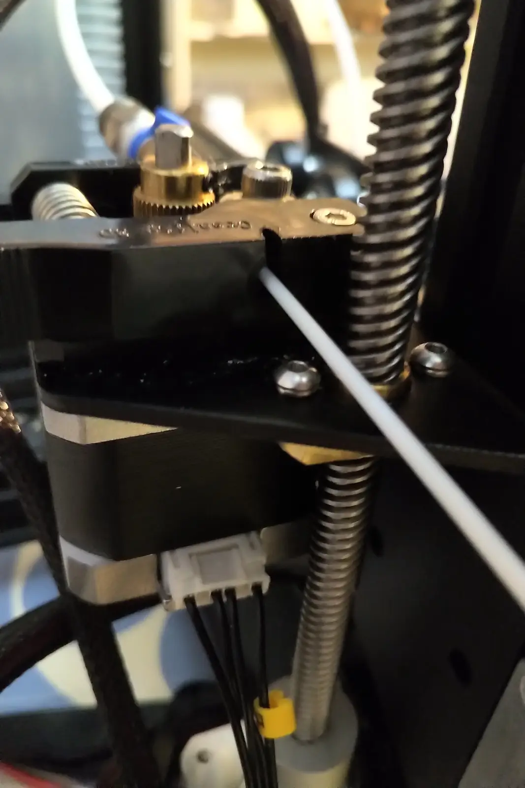 A close-up photo of a 3D printer's extruder mechanism, showing a white filament feeding through a black metal assembly with…