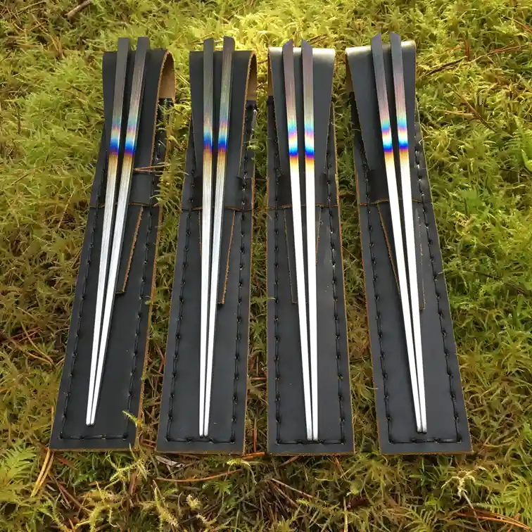 Four sets of chopsticks with iridescent rainbow-colored tips are neatly arranged in black leather cases on a bed of green…