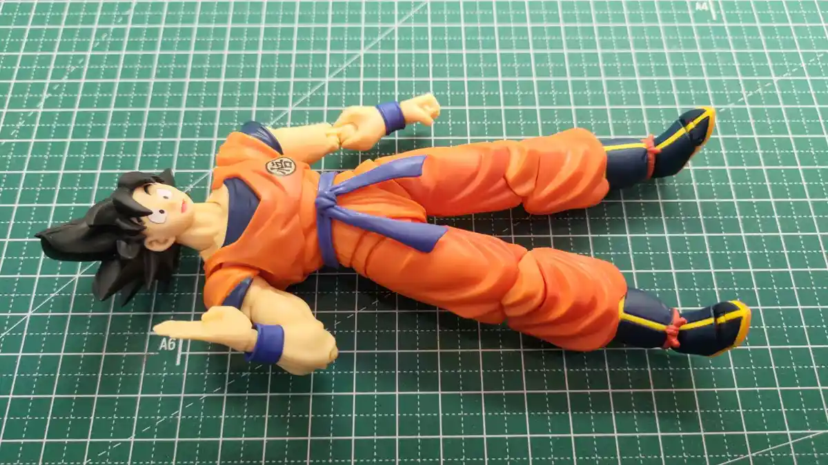 A plastic action figure of the anime character Goku from Dragon Ball, lying flat on his back on a green cutting mat with…