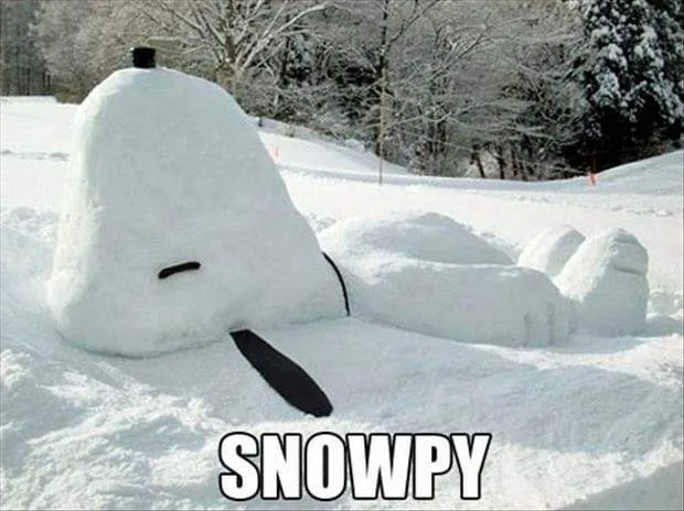 A snow sculpture shaped like the cartoon character Snoopy, lying on its back in a snowy landscape with trees in the…