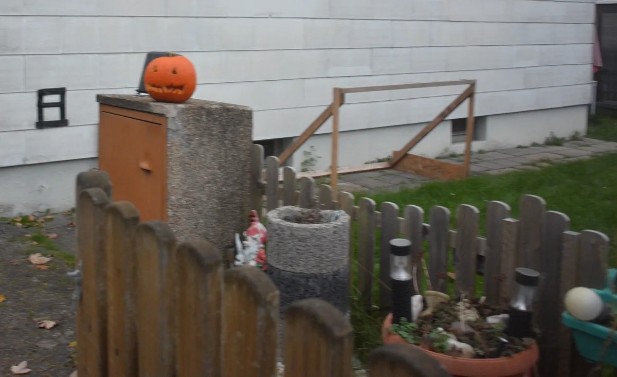 A carved orange jack-o'-lantern with triangular eyes and a jagged grin sits atop a concrete utility box in a backyard. The…