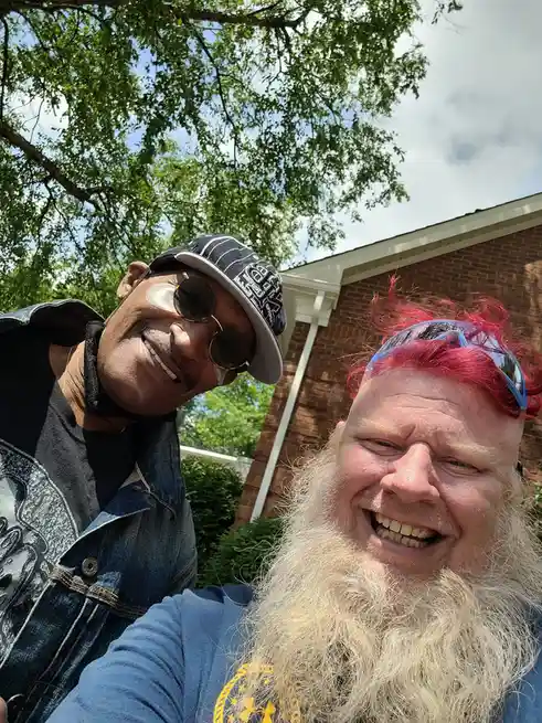 Two men are taking a selfie outdoors in front of a brick house with trees in the background. The man on the left wears a…