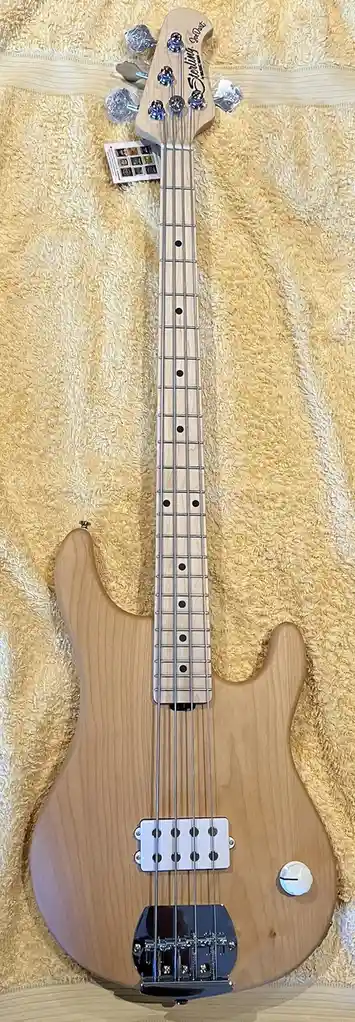 A light-wood, four-string electric bass guitar with a natural finish lies on a yellow terry cloth towel. The headstock has…
