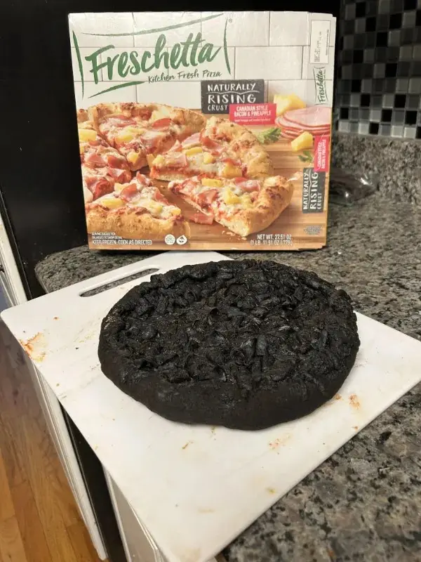 A burnt black pizza sits on a white cutting board in front of its Freschetta frozen pizza box, which shows a colorful…
