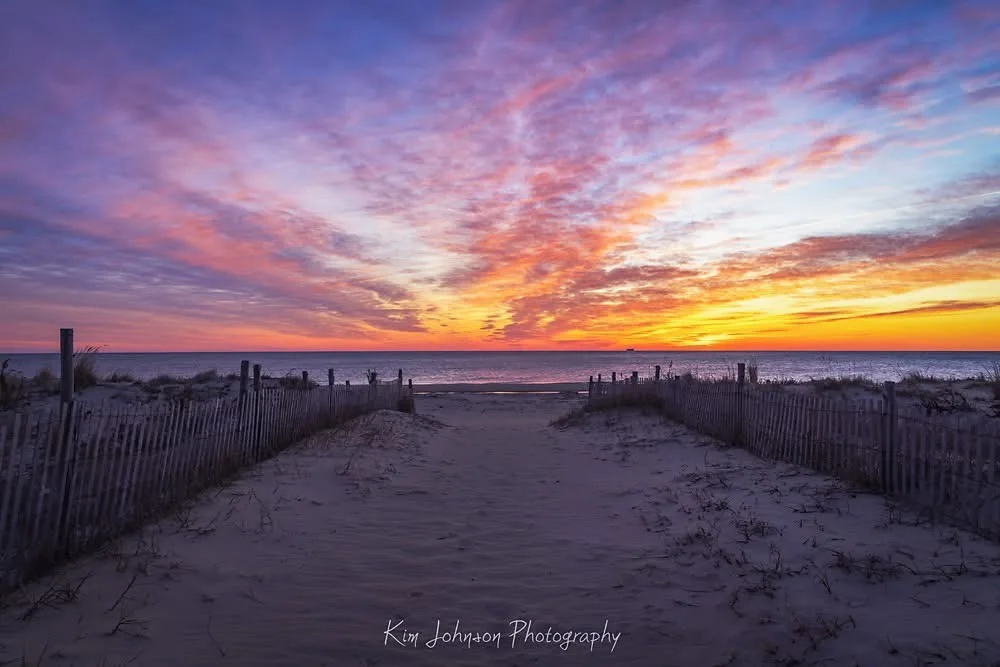 A serene beach sunset photo with a wooden fence-lined path leading to the ocean, featuring vibrant orange, pink, and purple…