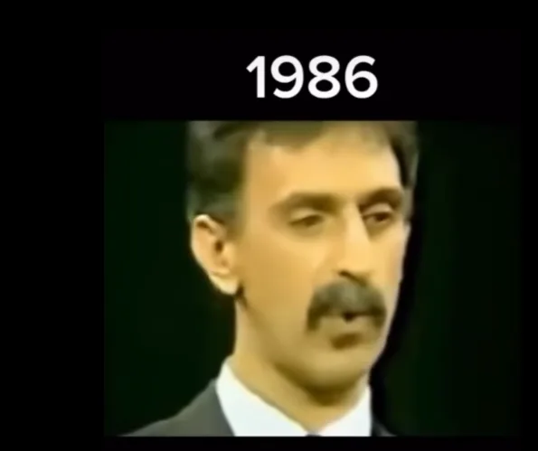 A grainy, low-resolution video still shows a man with a mustache and dark hair wearing a suit and tie, looking slightly to…