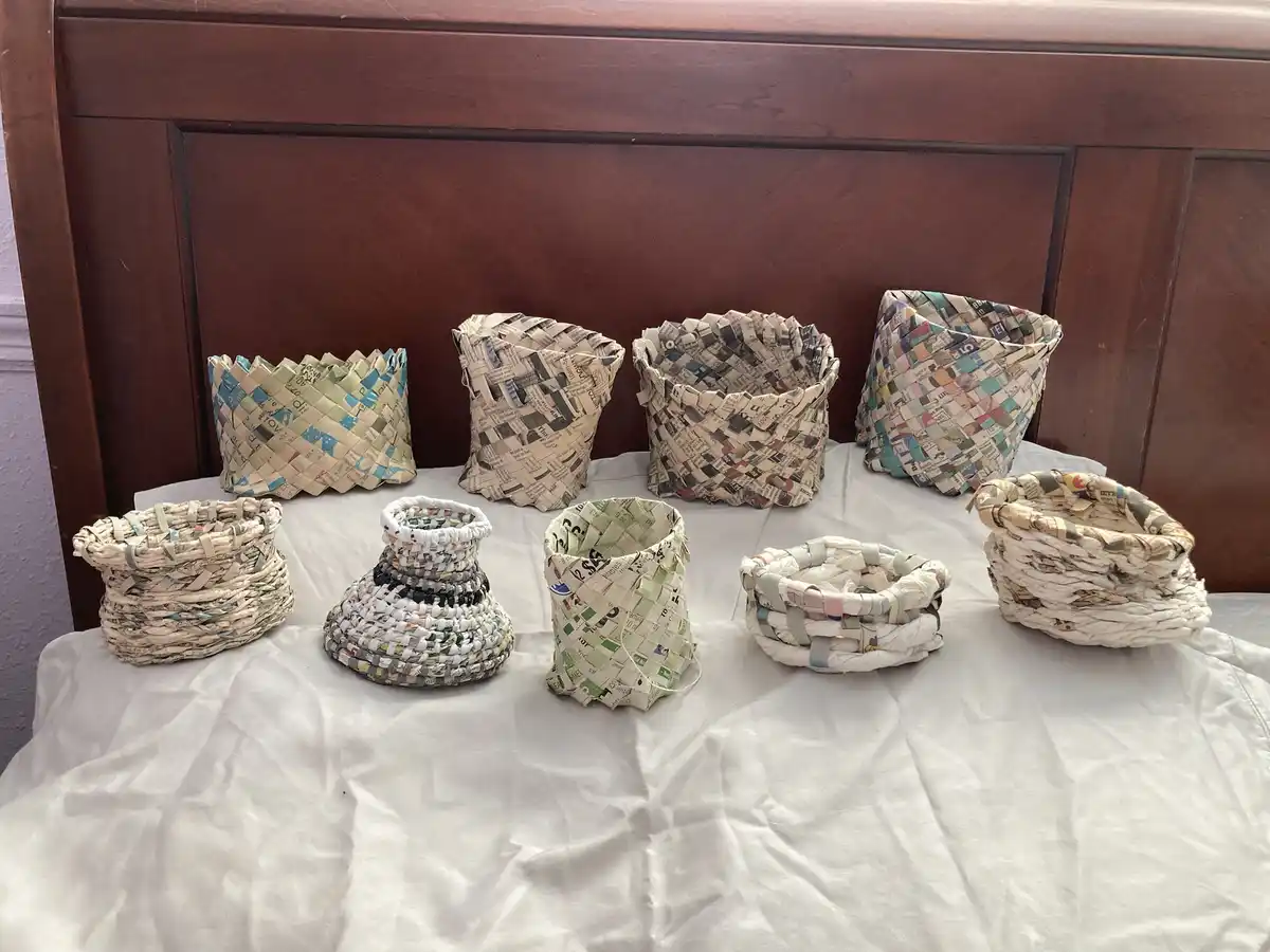 A collection of nine handmade baskets, woven from strips of newspaper with various patterns and colors, are arranged on a…
