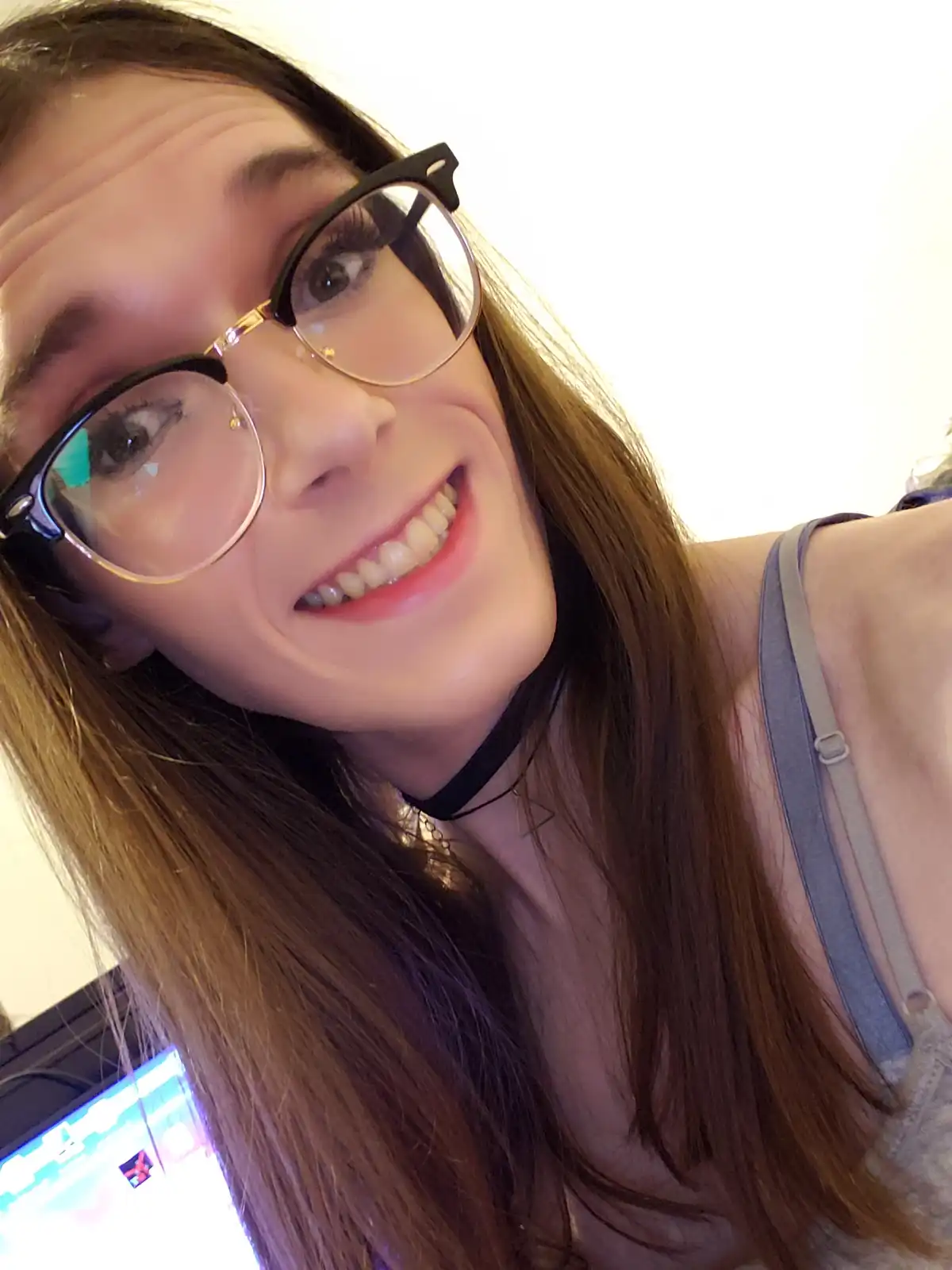 A selfie of a young woman with long brown hair, wearing black-rimmed glasses and a grey tank top, smiling broadly at the…