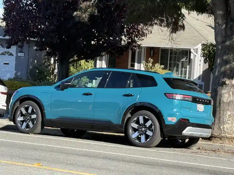 A bright turquoise Honda Prologue electric SUV is parked on a residential street, angled to show its side profile and rear…