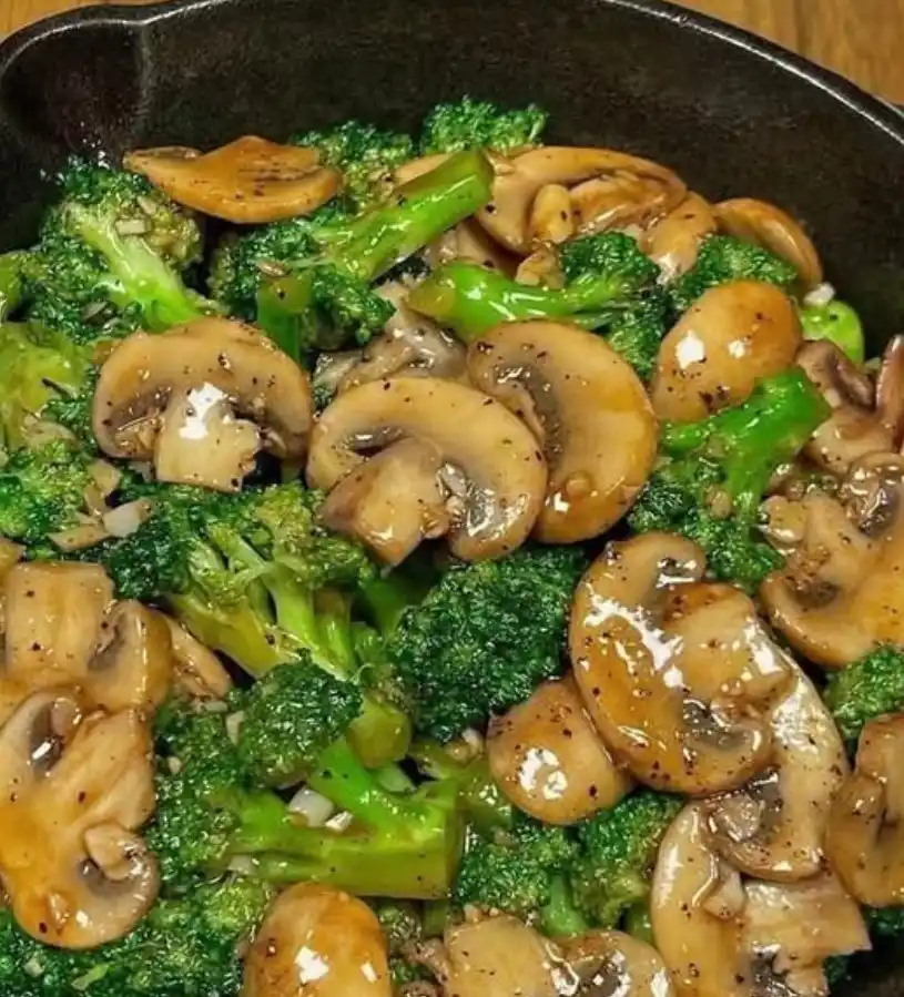 A close-up photo of sautéed broccoli florets and sliced mushrooms in a dark skillet, coated in a glossy sauce and sprinkled…