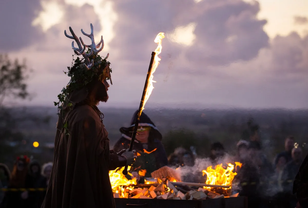 A person in a dark robe and antlered headpiece holds a flaming torch at a bonfire event, with other costumed attendees…