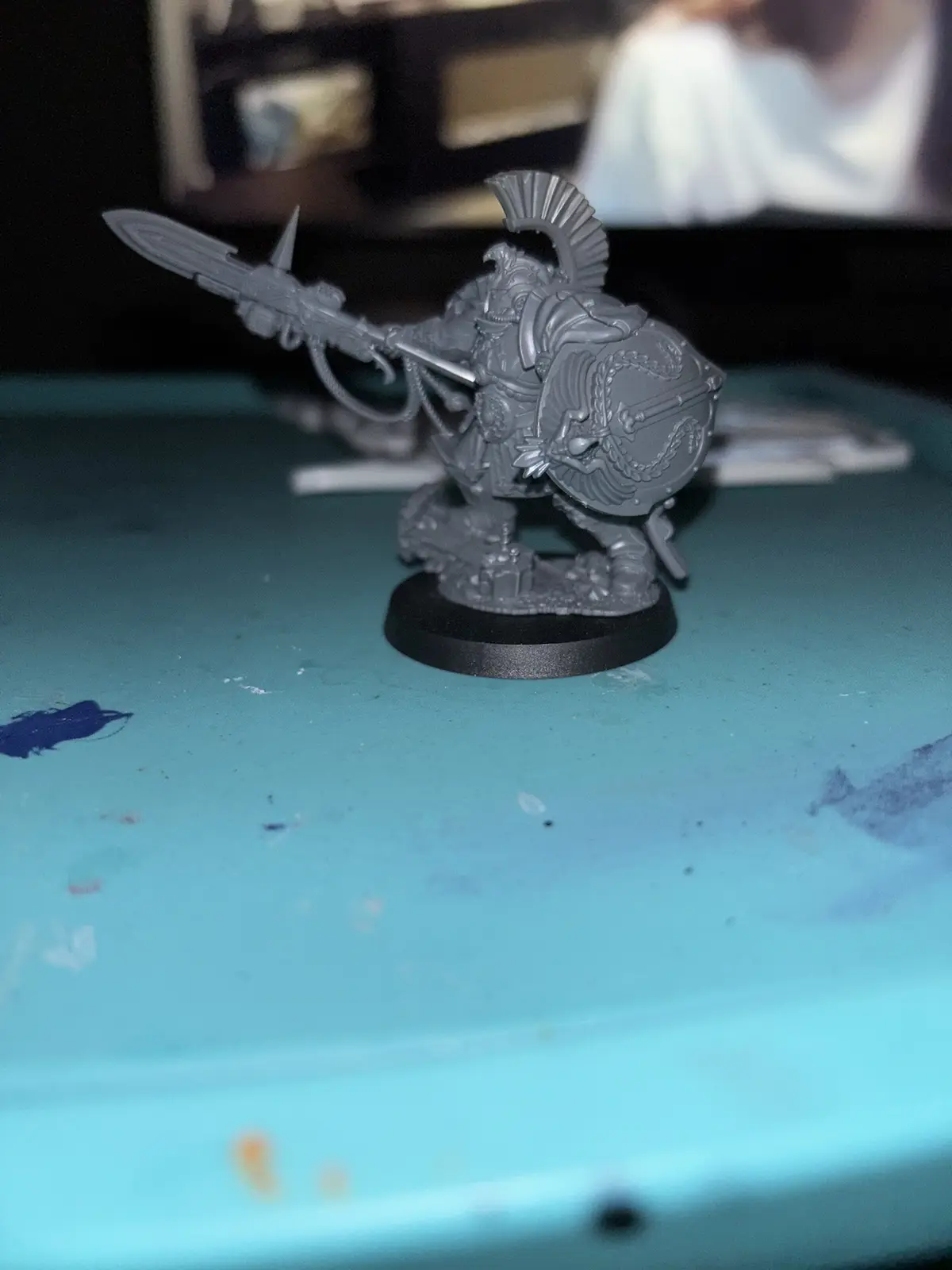 A gray, unpainted fantasy warrior miniature stands on a black base atop a teal surface with paint splatters. The figure…