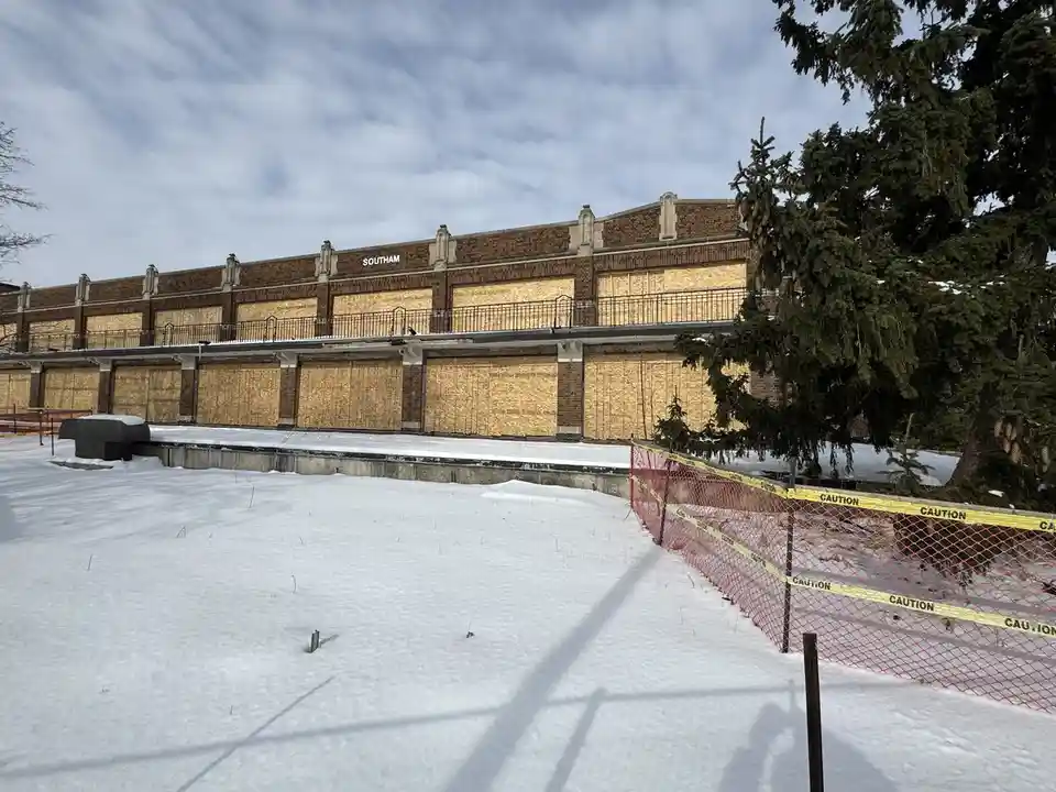 A snowy outdoor scene shows a large brick building with all windows boarded up with plywood. The building has a sign reading…