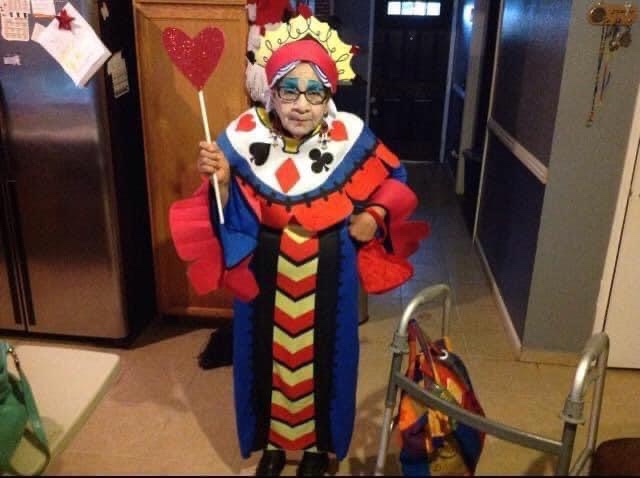An elderly woman wearing a colorful, elaborate jester or queen costume with a crown, holding a red heart-shaped wand, stands…