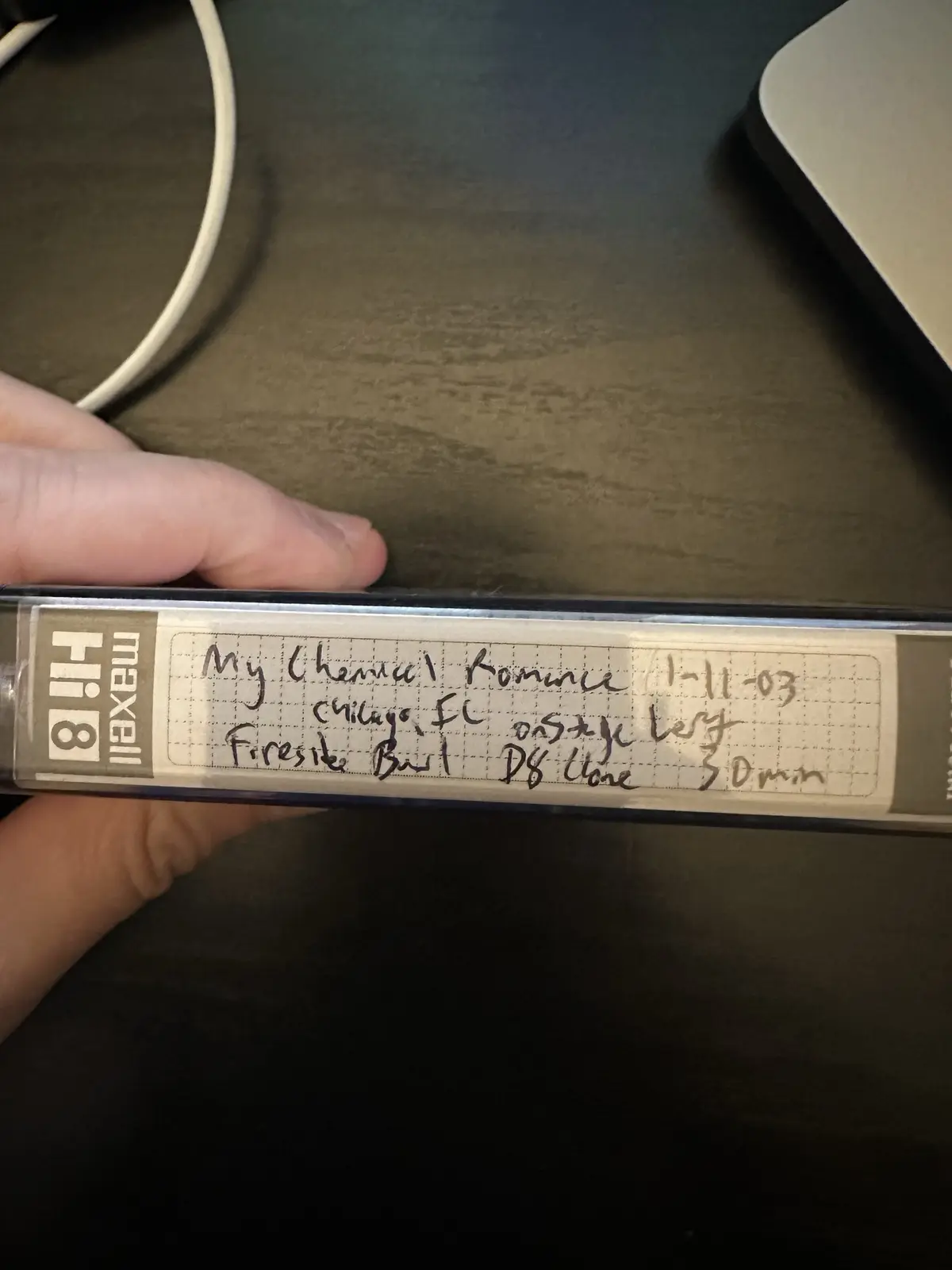 A hand holds a Maxell Hi8 video cassette tape, showing a handwritten label that reads 'My Chemical Romance 1-11-03 chicago…