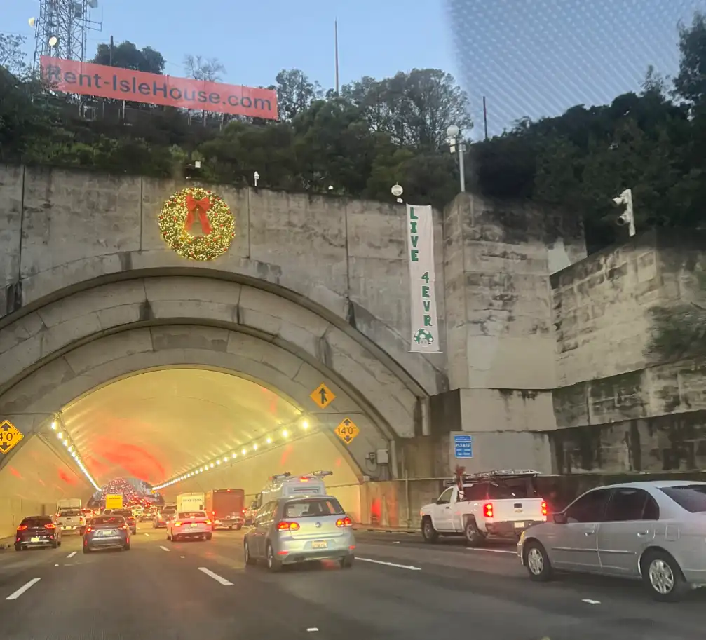 A photo of a highway tunnel entrance with cars in traffic, featuring a large Christmas wreath with a red bow on the concrete…