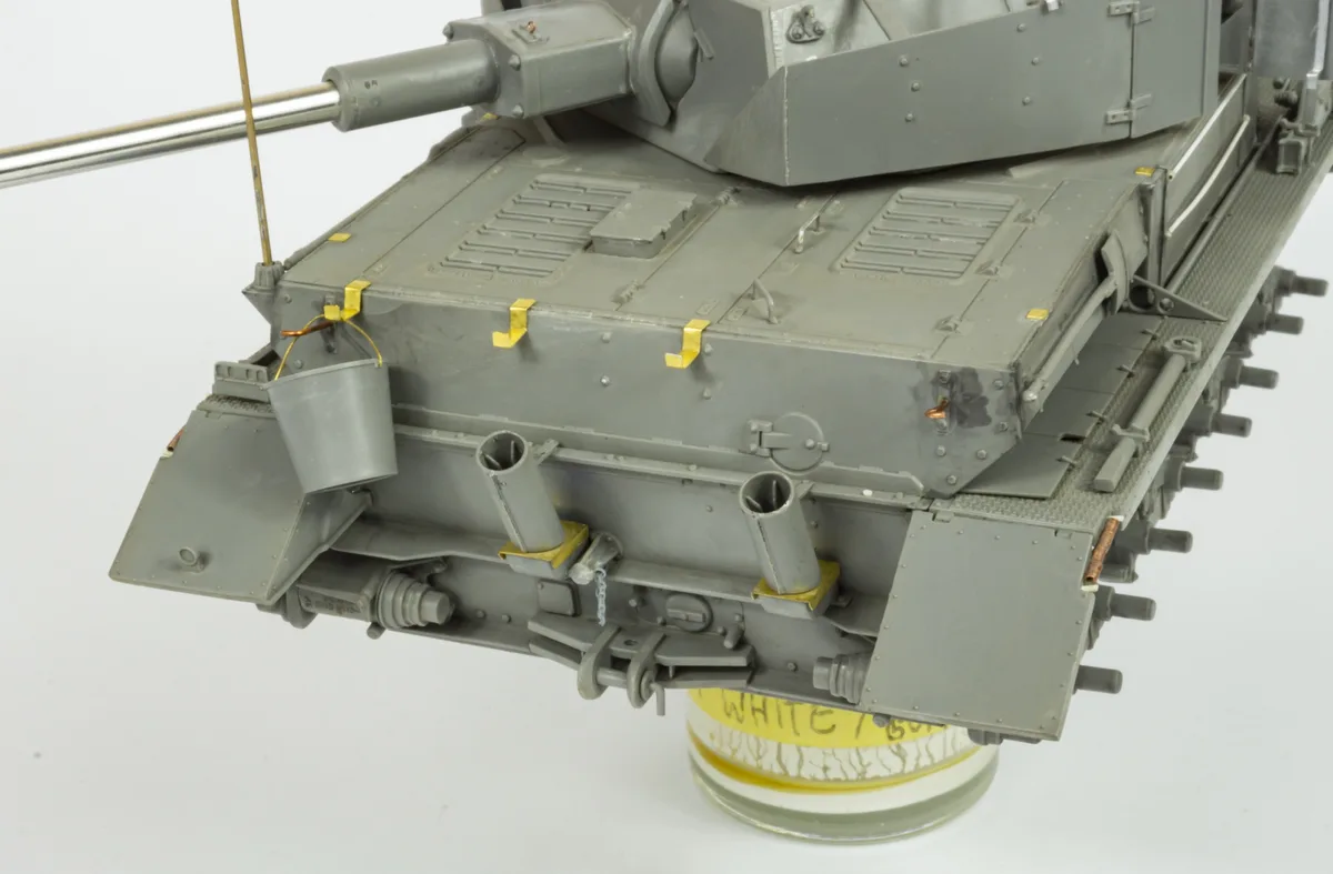 A close-up of a grey scale model tank with a small bucket hanging from its side, attached by yellow tape, and a yellow label…