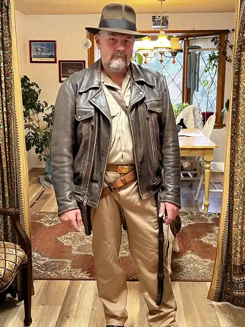 A man with a mustache and goatee stands in a home wearing a brown fedora, a brown leather jacket, beige pants, and a belt…