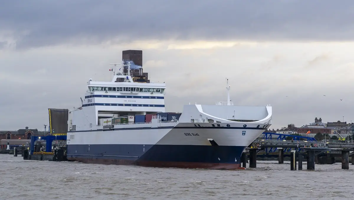 A large white and blue ferry named 'BORE SONG' is docked at a port under a cloudy sky, with industrial buildings and a…