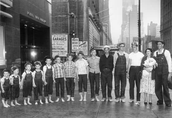 A black-and-white photo shows a family group, including several children in overalls, standing on a city street. The adults…