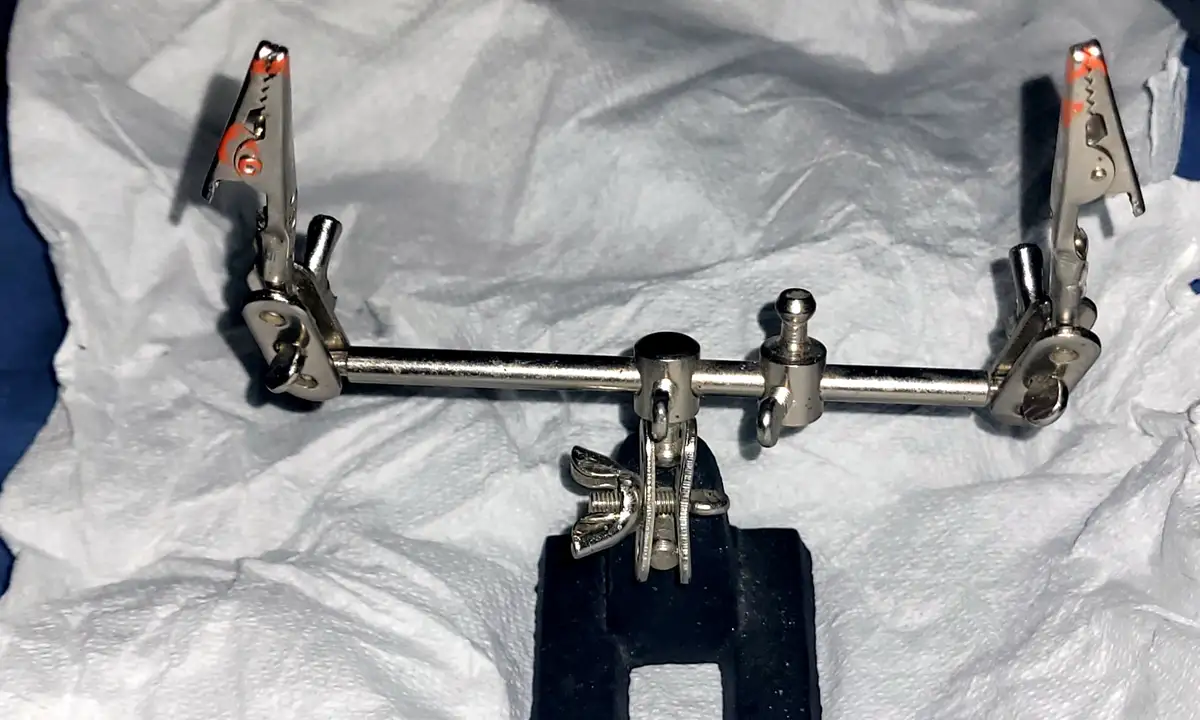 A metal contraption with two alligator clips at each end, mounted on a horizontal bar and secured to a black clamp base…