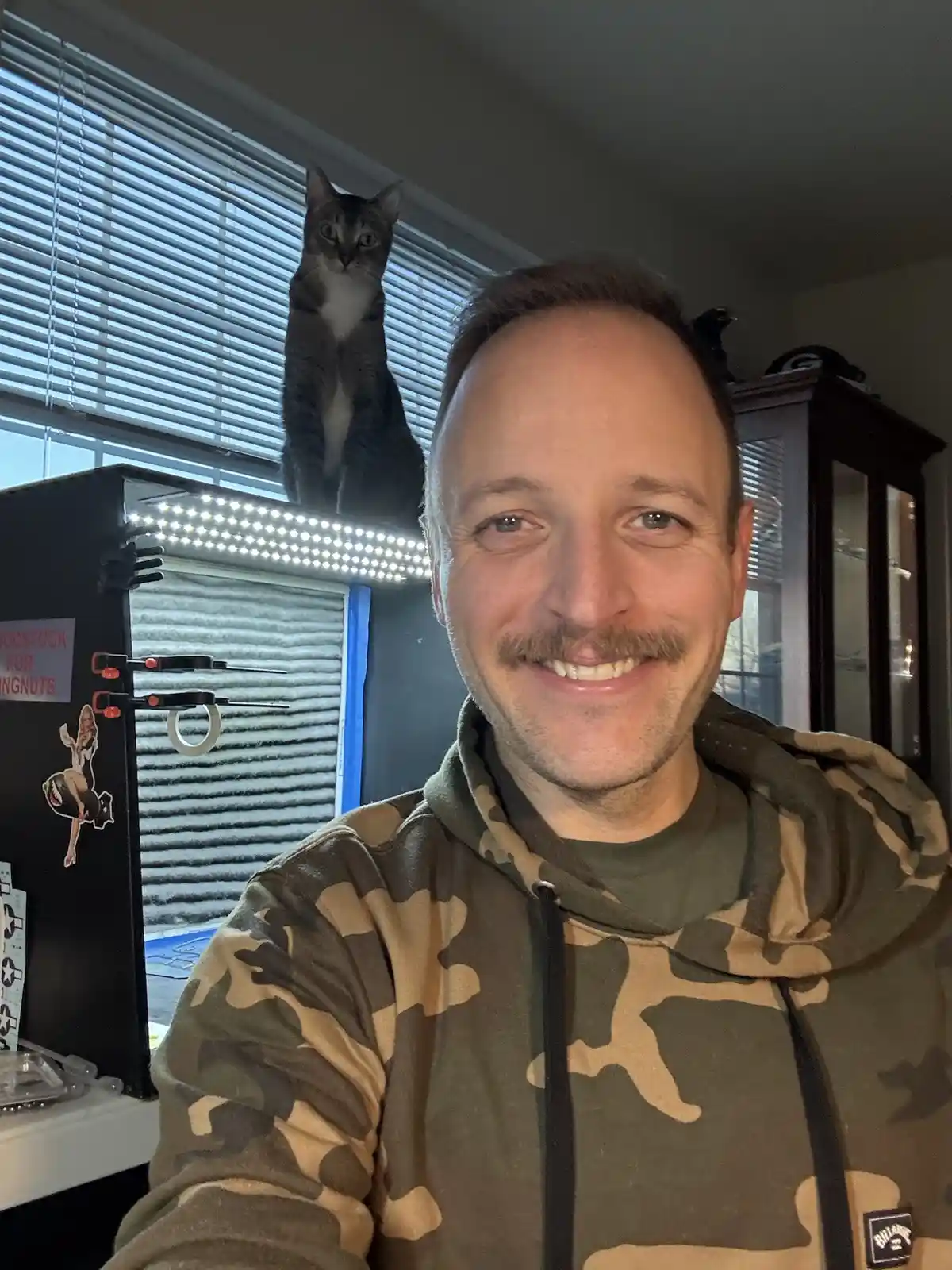 A man in a camouflage hoodie smiles at the camera while a grey tabby cat sits upright on top of a lighted box behind him…
