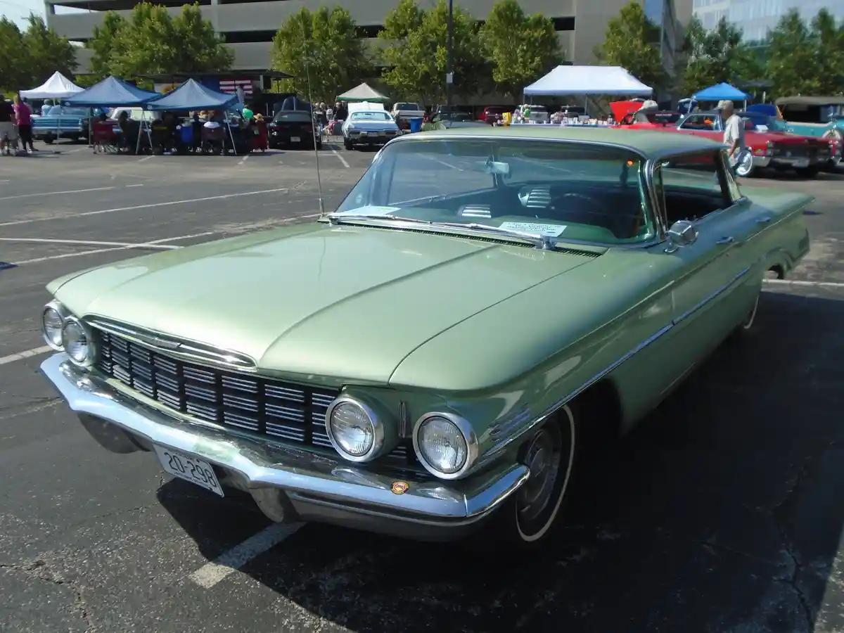 A light green 1950s classic car, likely a Chrysler, is parked at an outdoor car show with other vintage vehicles and tents…